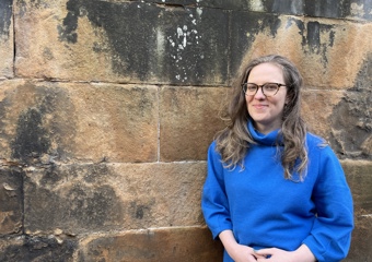 women, wearing a blue jumper and black rimmed glasses, standing in front of a wall and smiling