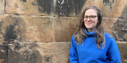 women, wearing a blue jumper and black rimmed glasses, standing in front of a wall and smiling